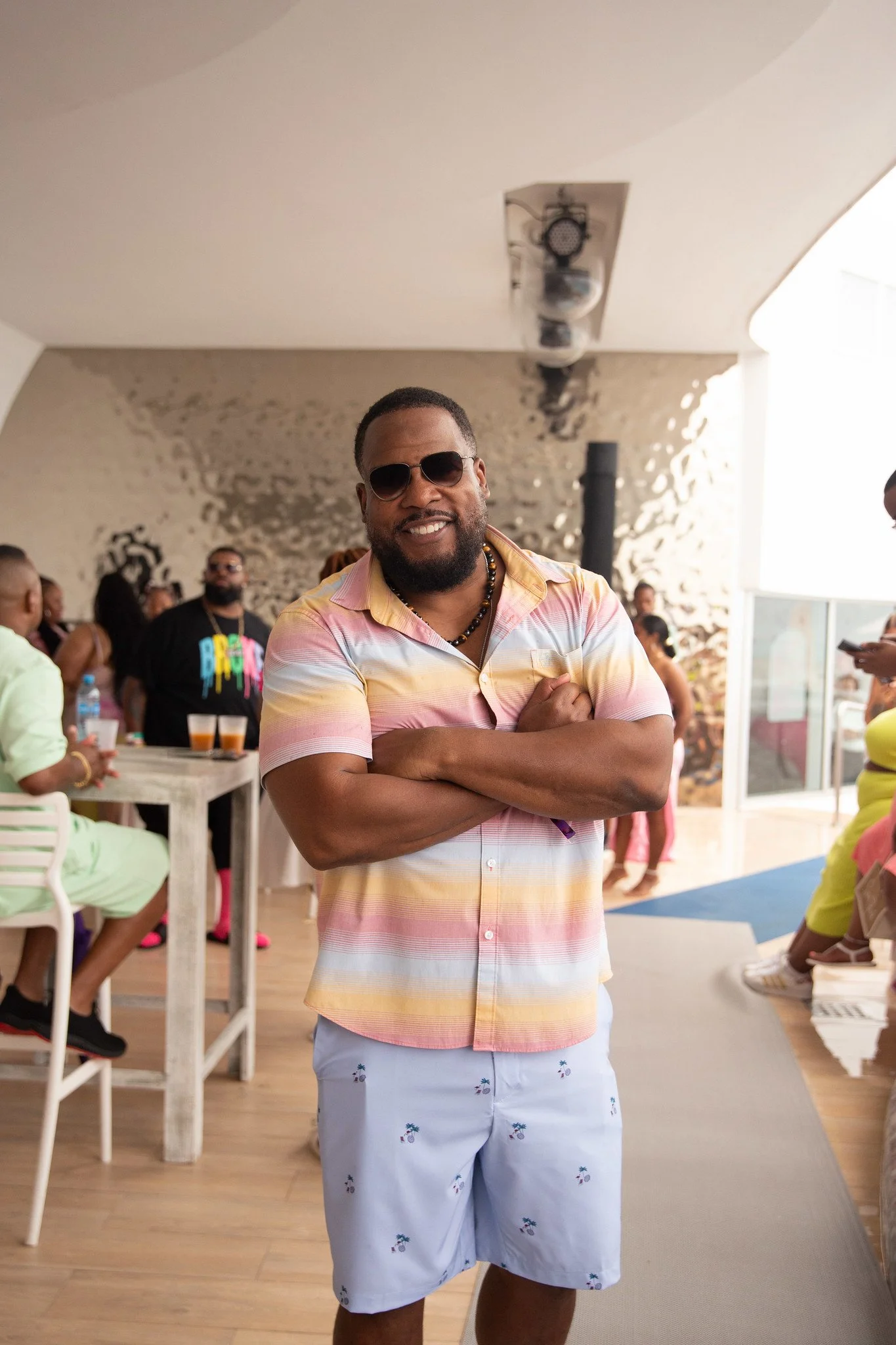 A man with sunglasses, a beard, and a colorful short-sleeved shirt stands with arms crossed at a social gathering or party.