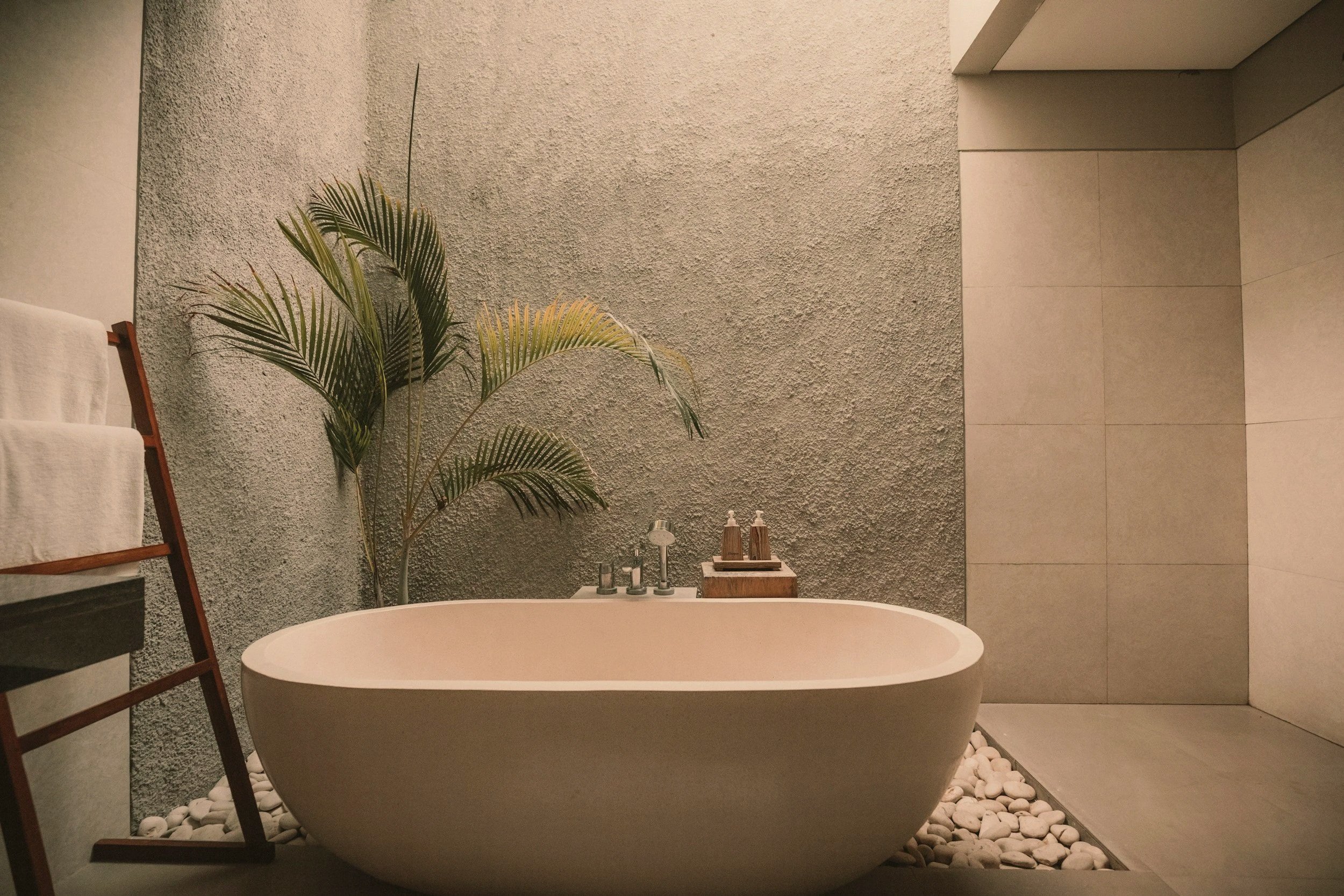 A modern bathroom with a standalone bathtub on white rocks, a textured gray wall, and a green potted plant. There is a wooden towel rack with white towels and a small wooden tray with toiletries.