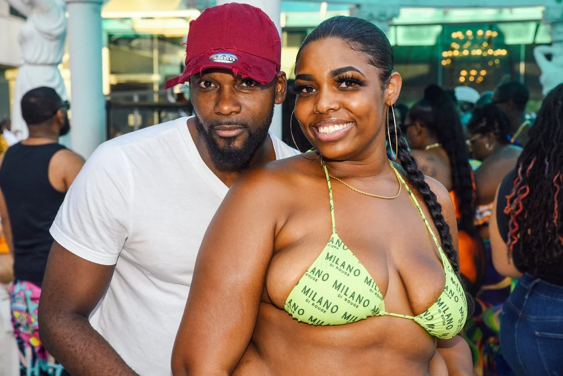 A man wearing a red cap and white t-shirt standing next to a smiling woman in a green bikini top with the word 'Milano' printed on it. They are at an outdoor event or party with other people in the background.