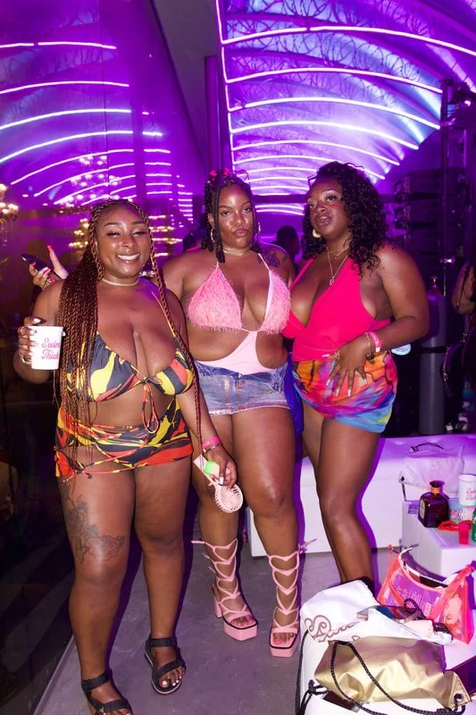Three women in colorful swimwear posing together at a party with purple neon lighting.