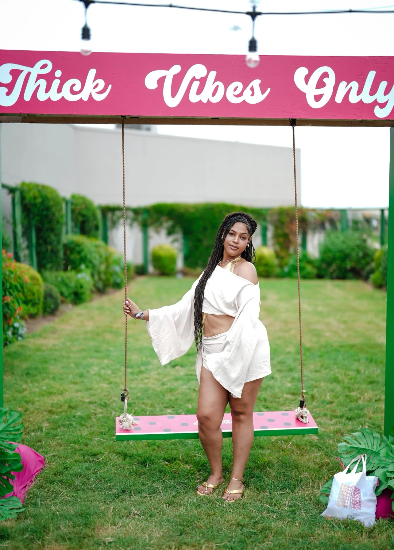 A woman with long, braided hair standing on a pink and green swing in a garden, wearing a white off-shoulder top and matching shorts, holding the swing rope, with a sign above that reads 'Thick Vibes Only'.