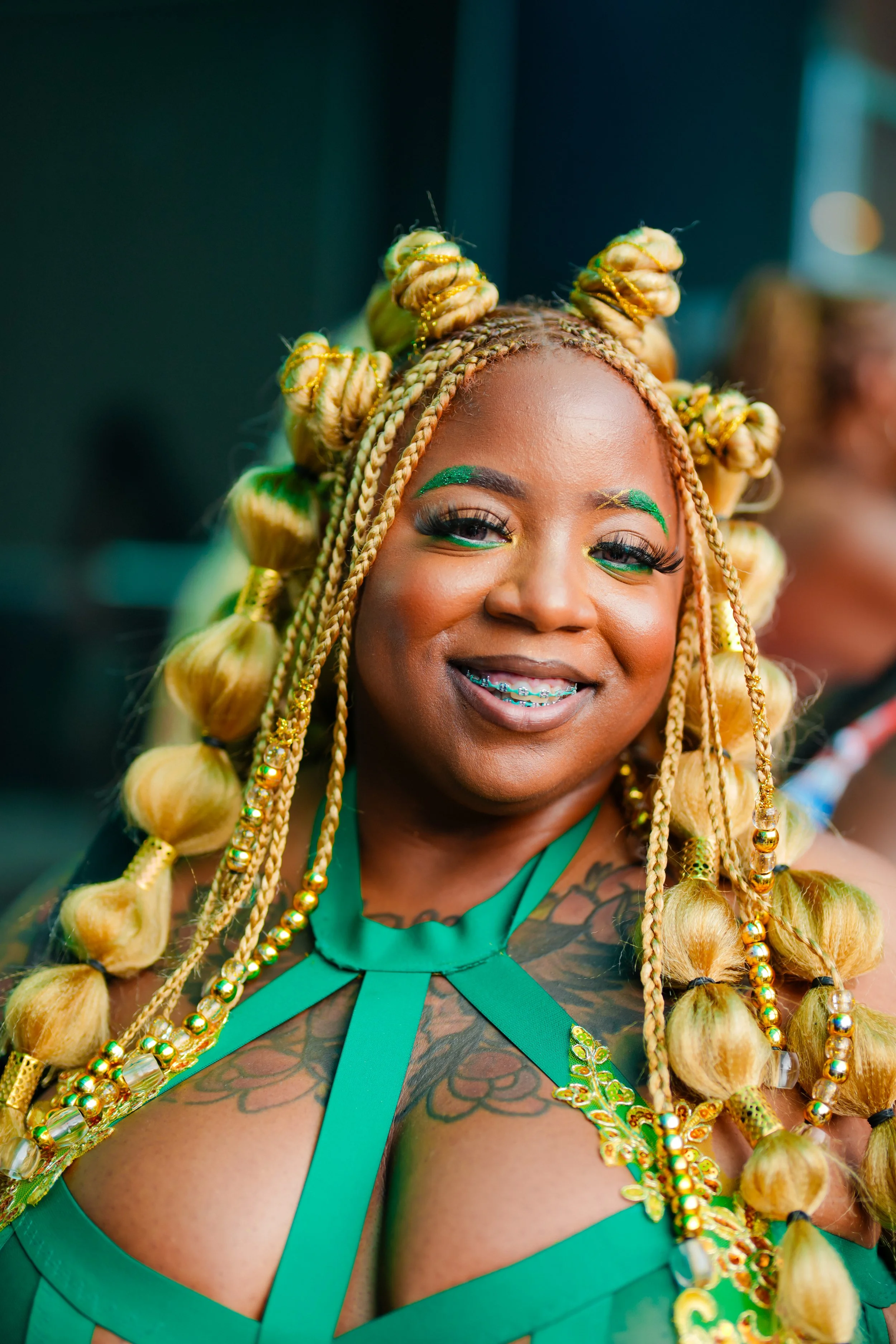 A smiling woman with braided, blonde hair styled in buns with beads, wearing green makeup and a green outfit with intricate details and tattoos on her chest.