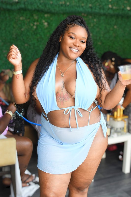 A woman in a light blue halter bikini top and skirt, smiling and holding a drink at a social gathering.