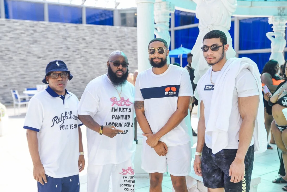 Four men standing together near a white sculpture at a poolside or outdoor venue, dressed casually in summer clothing and sunglasses.