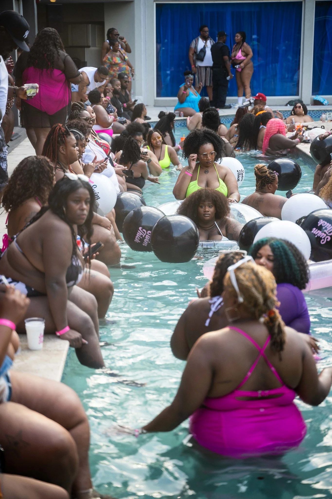 A crowded pool party with many women in swimsuits, some holding drinks and balloons, surrounded by onlookers and photographers.