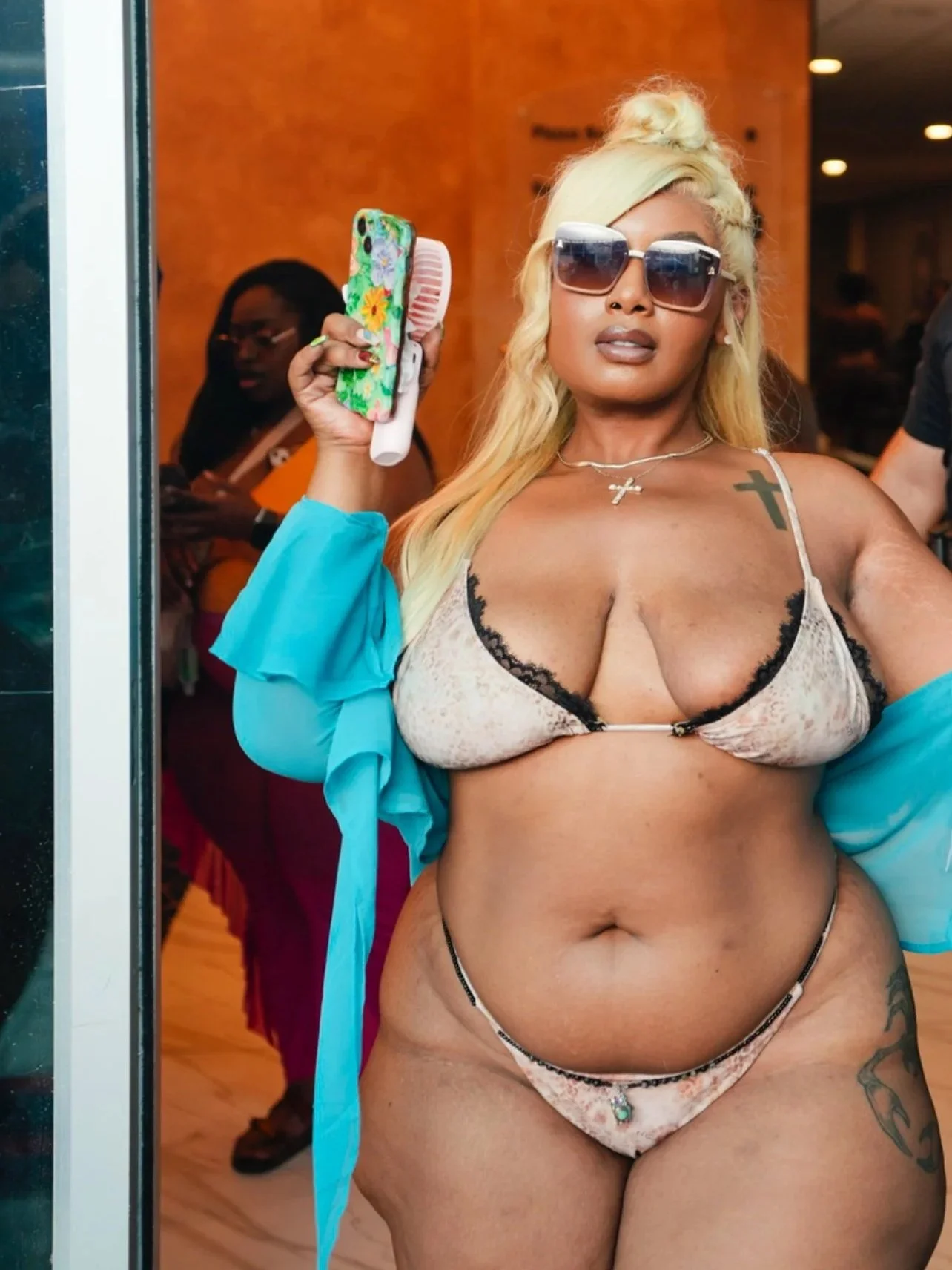 A woman with blonde hair, sunglasses, and tattoos on her shoulders and thigh, wearing a bikini and a blue cover-up, holding a phone with a floral case, standing in front of a mirror.