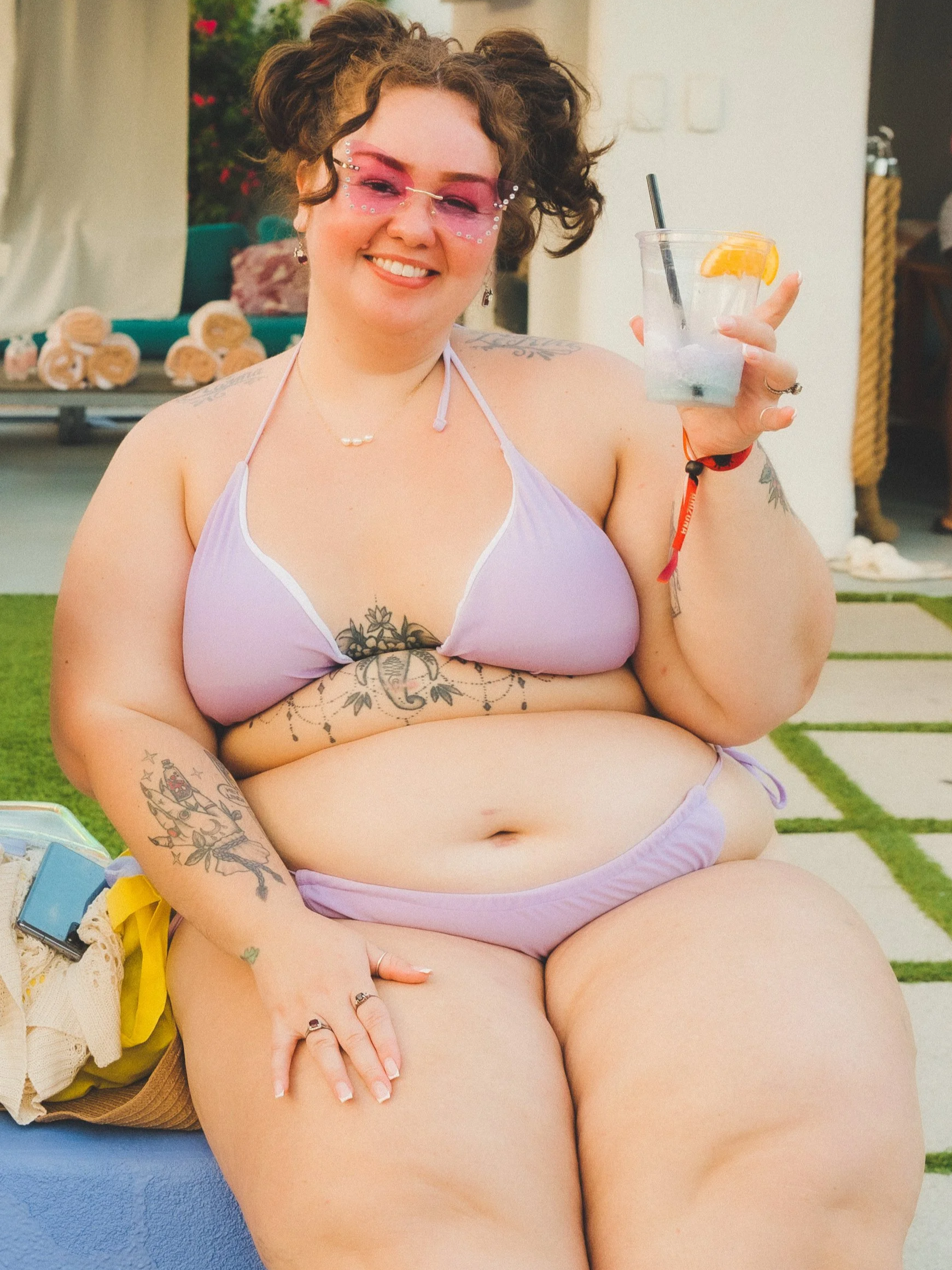 A woman in a lavender bikini holding a cup with a straw, outdoors at a pool or backyard setting, smiling at the camera.