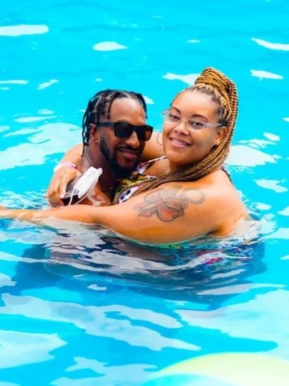A man and woman smiling and hugging in a swimming pool, with the woman having braids and a tattoo on her arm.