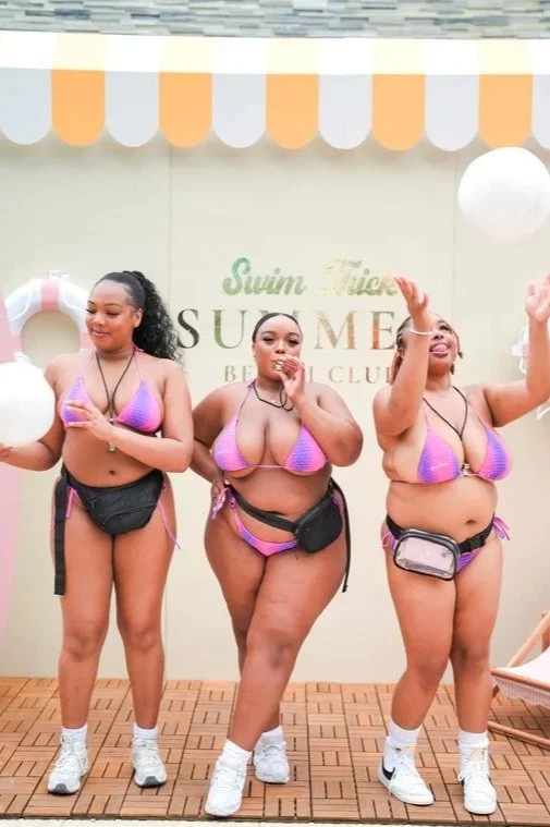 Three women in bikinis and fanny packs standing on a pool deck, celebrating at a summer-themed event with a backdrop reading 'Swim Classic Summer'.