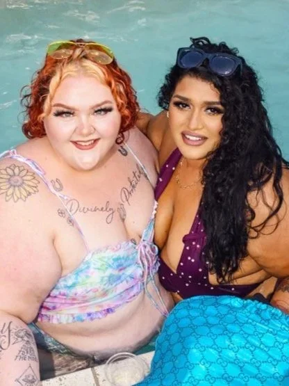 Two women in a swimming pool, smiling at the camera. One has curly red hair and a colorful swimsuit, the other has dark curly hair with sunglasses on her head and a purple swimsuit. They are close together, enjoying a sunny day.
