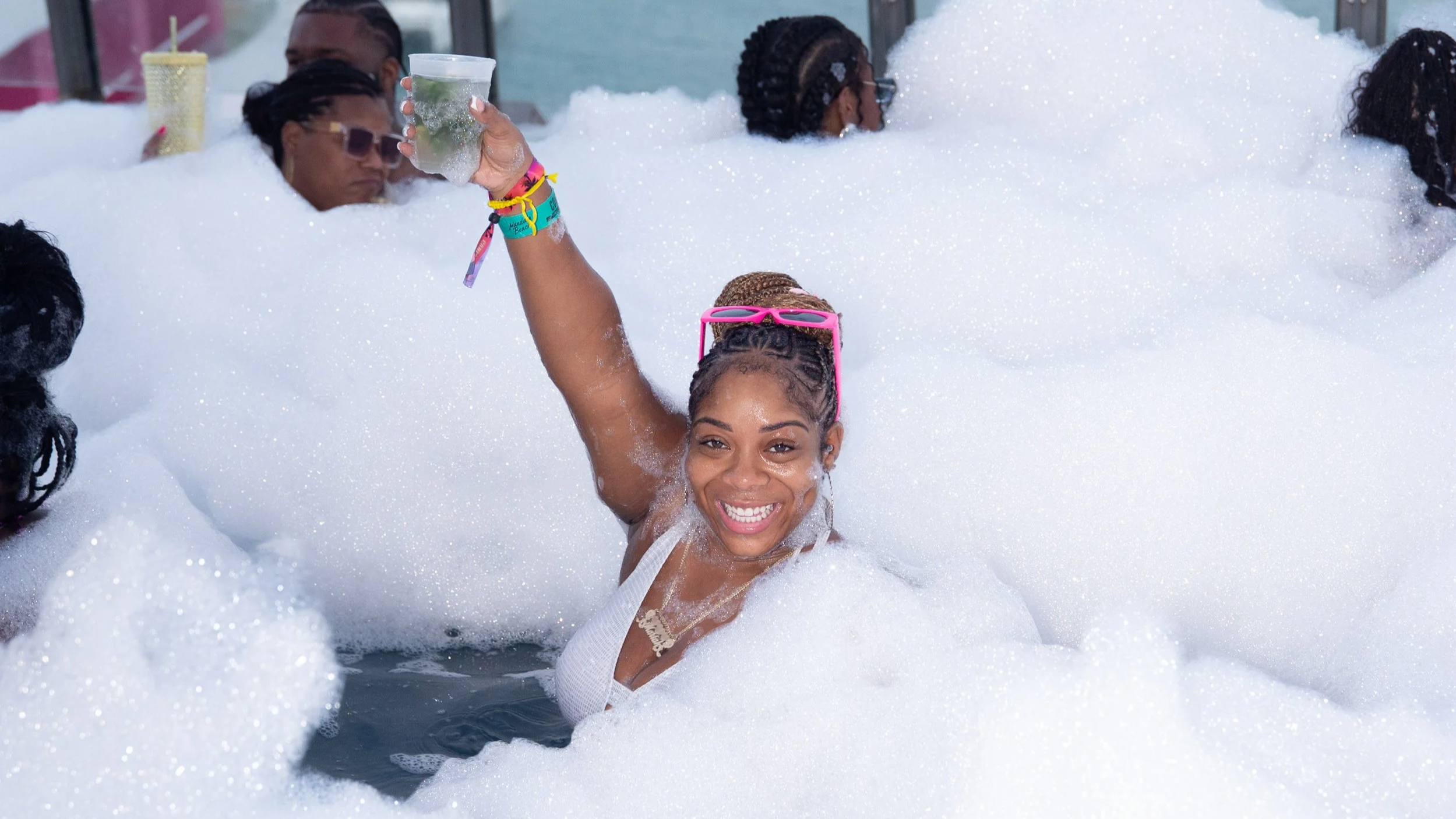 A group of people enjoying a bubble bath in a pool with foam. A smiling woman in the center is raising a drink, wearing pink sunglasses and a white top, surrounded by others relaxing in the foam.