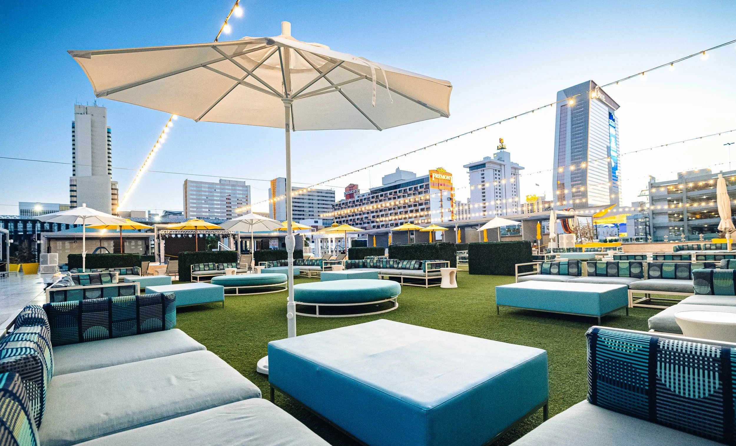 Urban rooftop lounge with outdoor seating, umbrellas, and string lights, overlooking city skyline with tall buildings in the background.