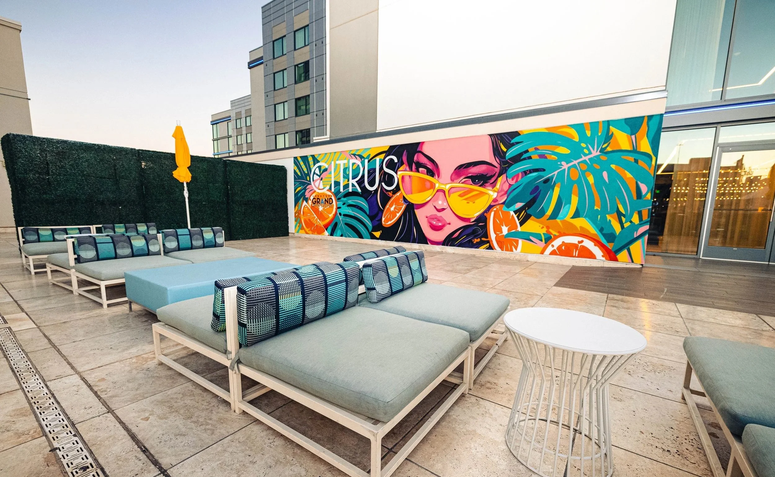 Outdoor rooftop lounge area with modern seating, a colorful mural featuring a woman with sunglasses and tropical fruit, and tall buildings in the background.