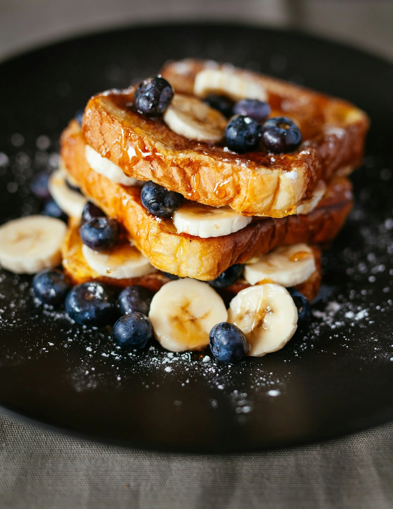 Stacked French toast with bananas, blueberries, and syrup on a black plate.