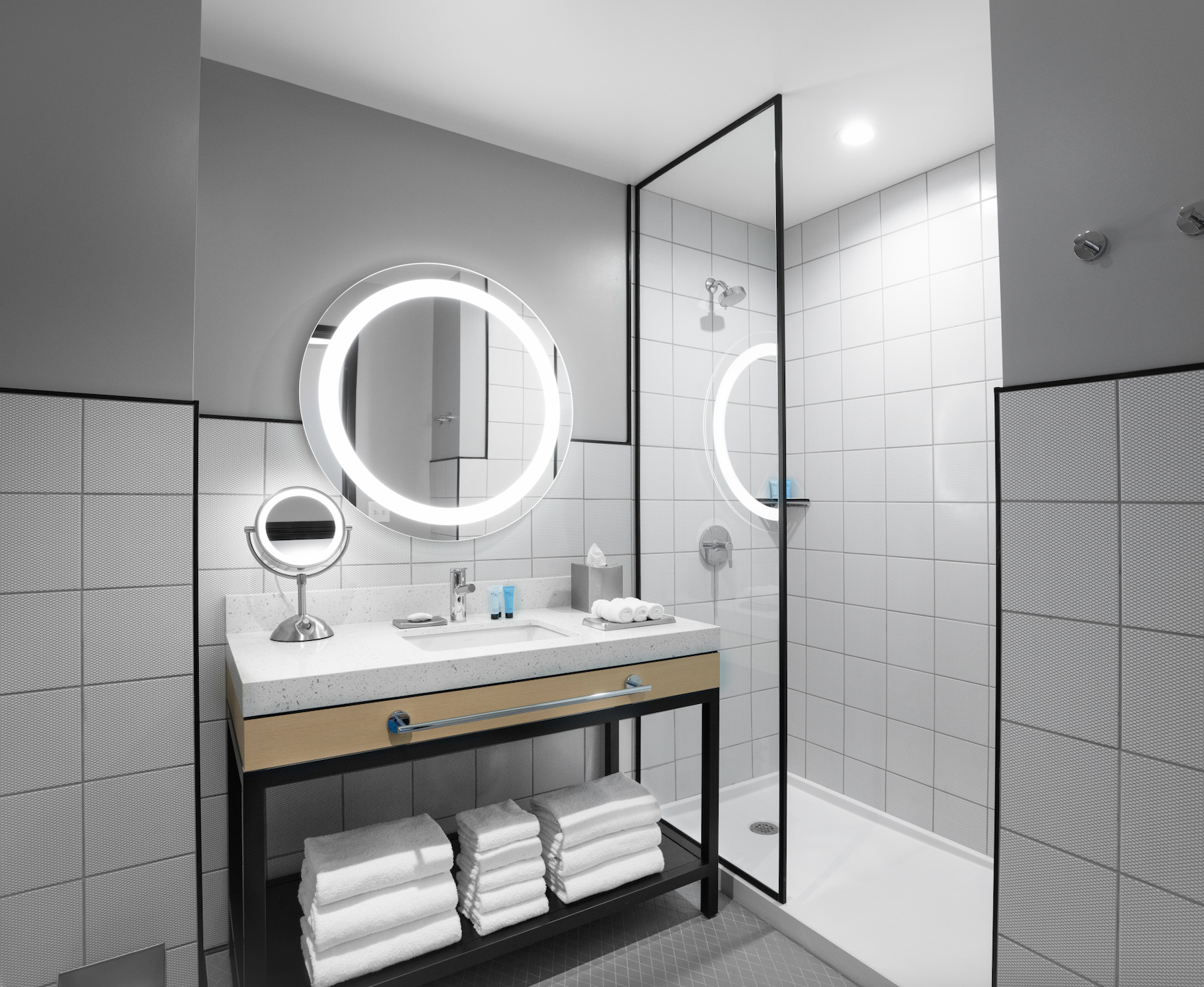 Modern bathroom with illuminated circular mirrors, a shower with glass enclosure, white tiled walls, a vanity with a sink, toiletries, and neatly folded towels.