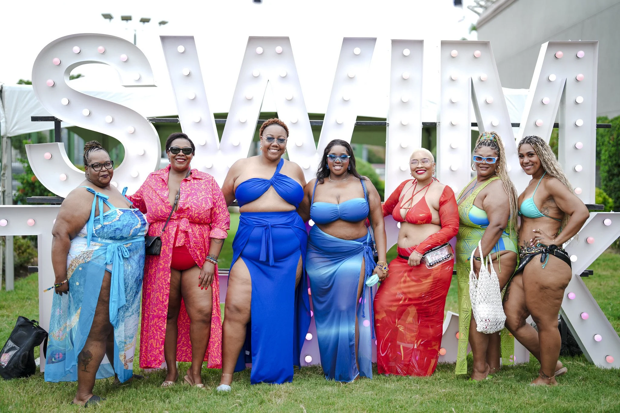 Group of eight women in colorful swimwear and cover-ups, smiling, standing in front of large illuminated "SAX” sign outdoors.