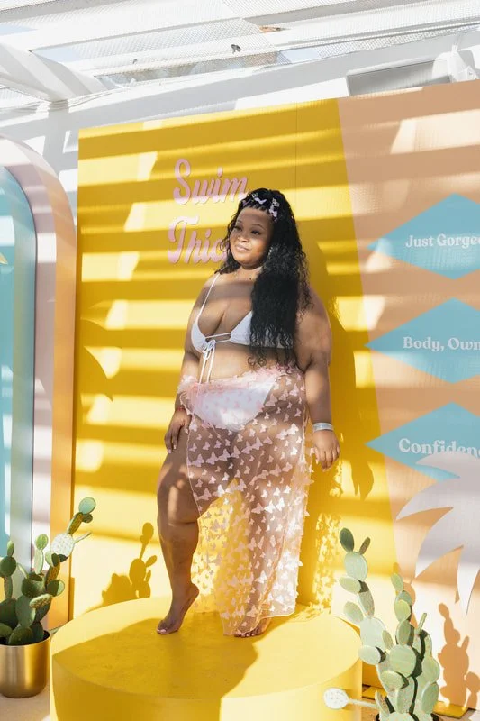 A woman stands on a yellow platform in front of a yellow backdrop with text, wearing a white bikini top and a sheer floral skirt. She has long curly black hair and is posing with one leg slightly forward. There are potted cacti around the platform, a