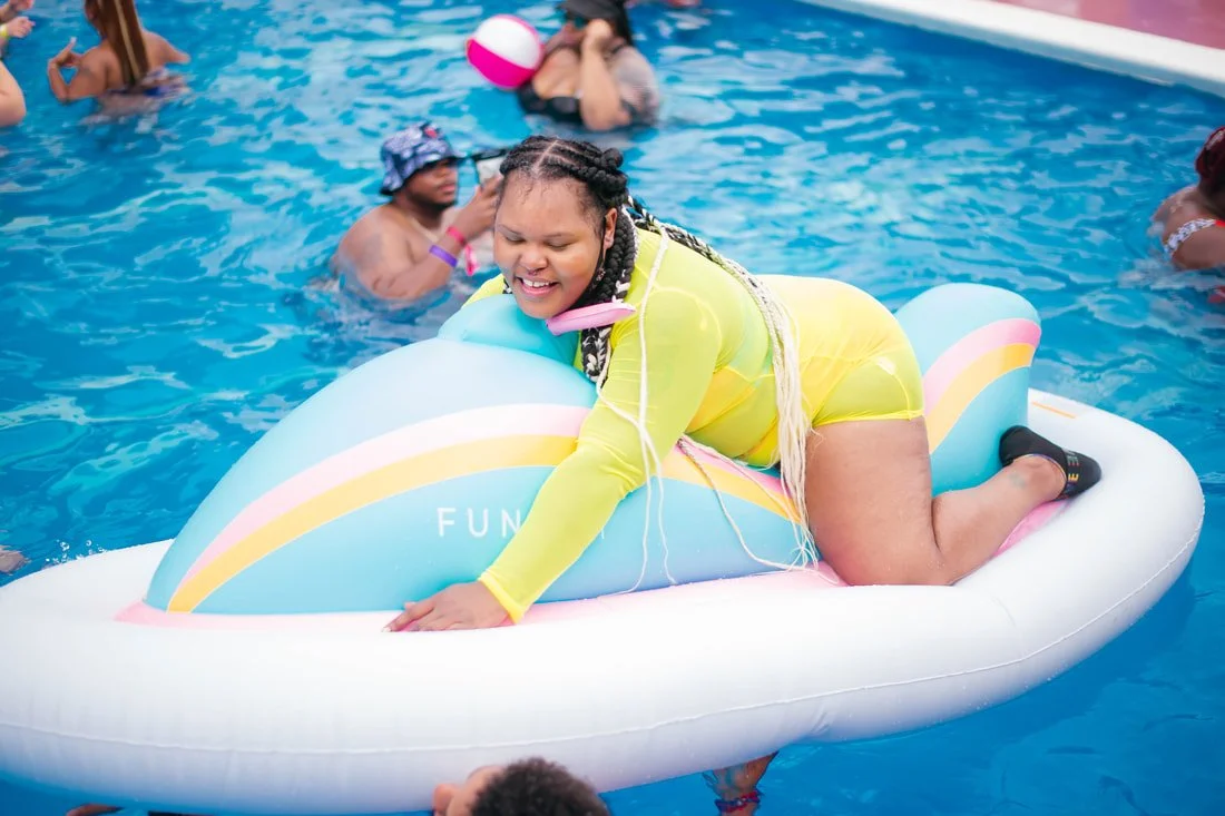 A young woman in a yellow swimsuit and hair braids is on an inflatable rainbow-colored float in a swimming pool, appearing to be enjoying herself with her eyes closed and a smile on her face, surrounded by other swimmers.