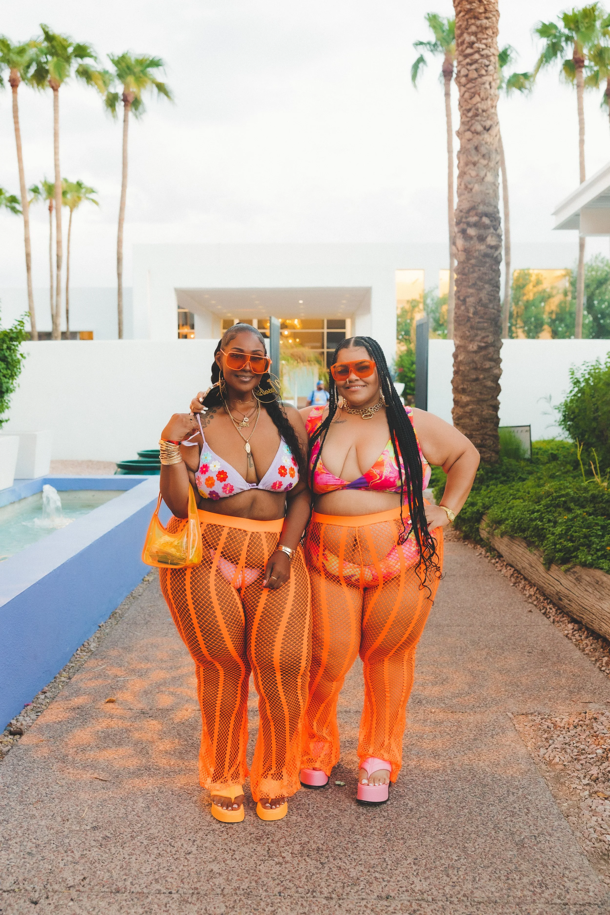 Two women in colorful swimwear and orange mesh pants standing outdoors near a small fountain, with palm trees and modern white buildings in the background.