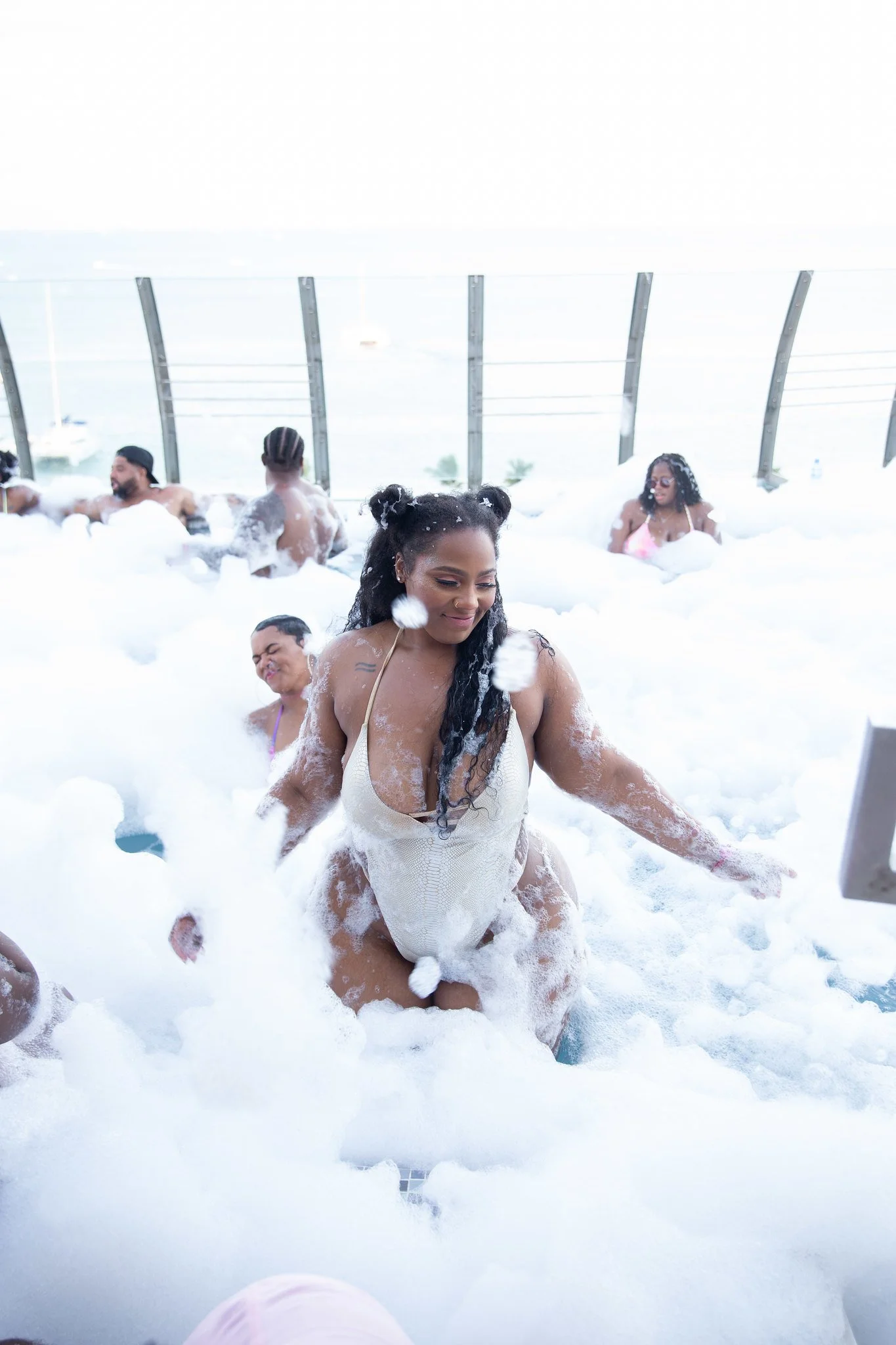 People enjoying a bubble bath on an outdoor rooftop with glass railings and city view.