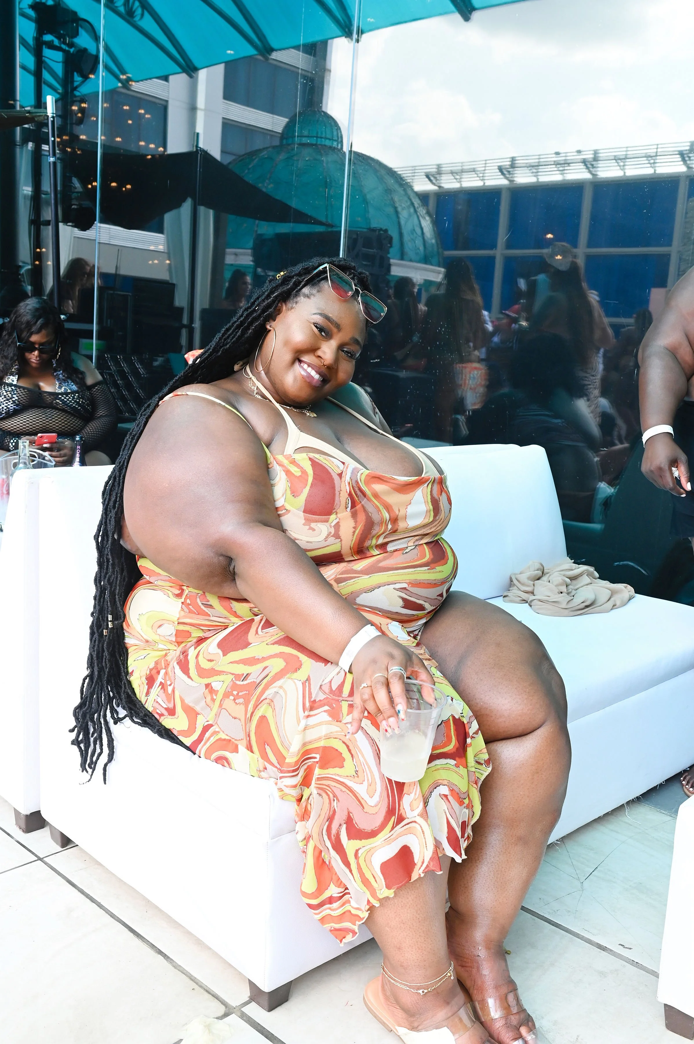 A woman with long dreadlocks and sunglasses sitting on a white sofa at a social event, holding a drink, smiling at the camera, wearing a colorful patterned dress.