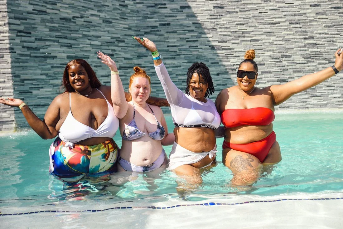 Four women in swimsuits enjoying a pool with a gray tiled wall in the background, smiling and raising their arms.