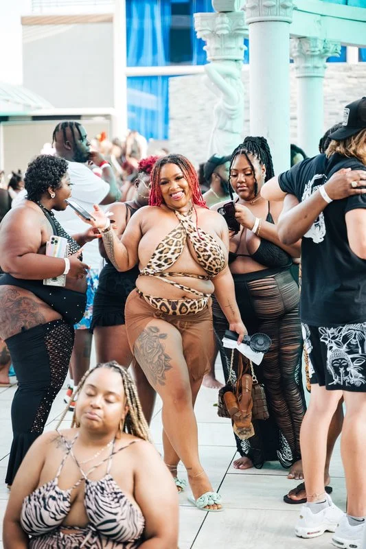 A group of women and a man at a pool party, some wearing swimwear and others in casual summer clothes, with a woman in a leopard-print bikini top and brown bottoms smiling and dancing in the center.