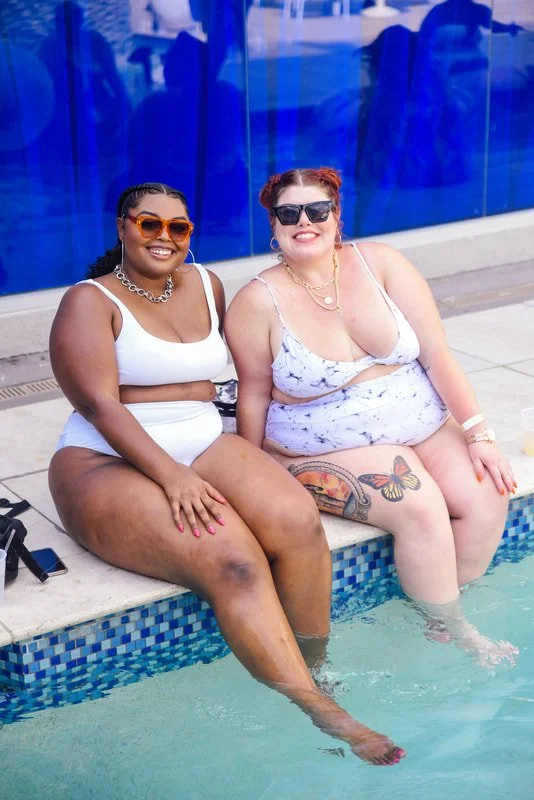Two women in swimsuits sitting poolside with their feet in the water, smiling at the camera, wearing sunglasses and jewelry.