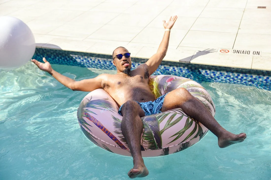 A man relaxing on a pink inflatable pool float with a tropical palm print, wearing sunglasses and swim trunks, in a swimming pool.