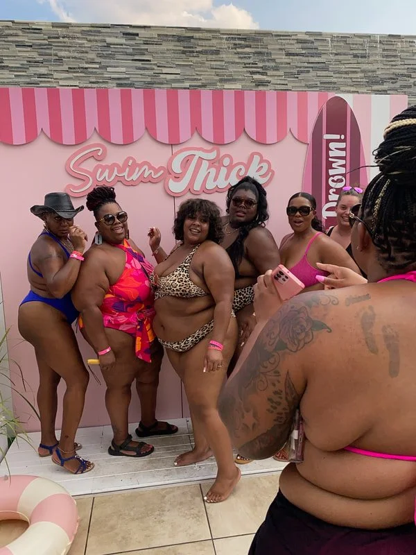 Group of women in swimsuits taking a photo at a poolside event labeled 'Swim Thick' with a pink and striped backdrop.