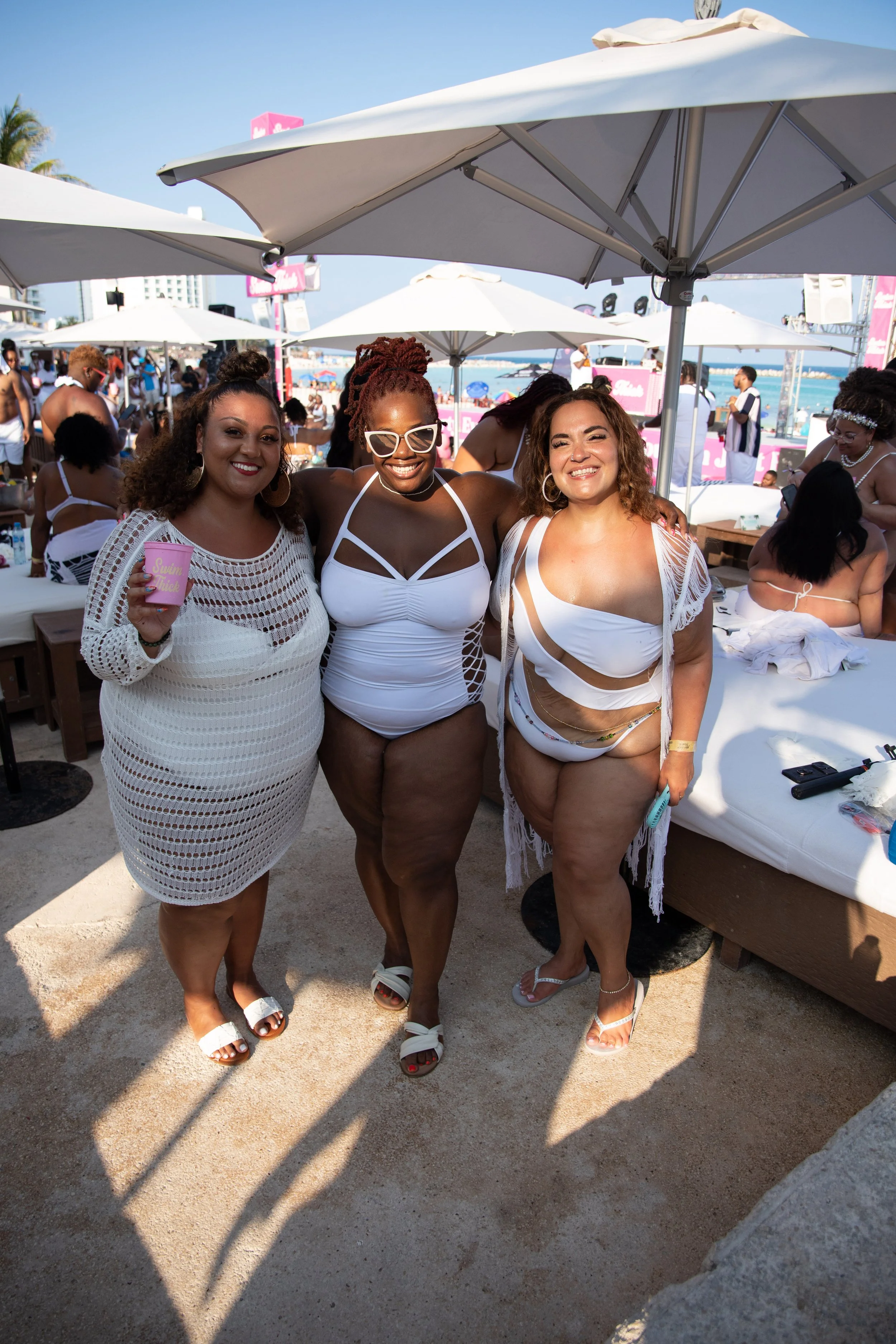 Three women in swimwear smiling and posing under beach umbrellas at a crowded beach event.