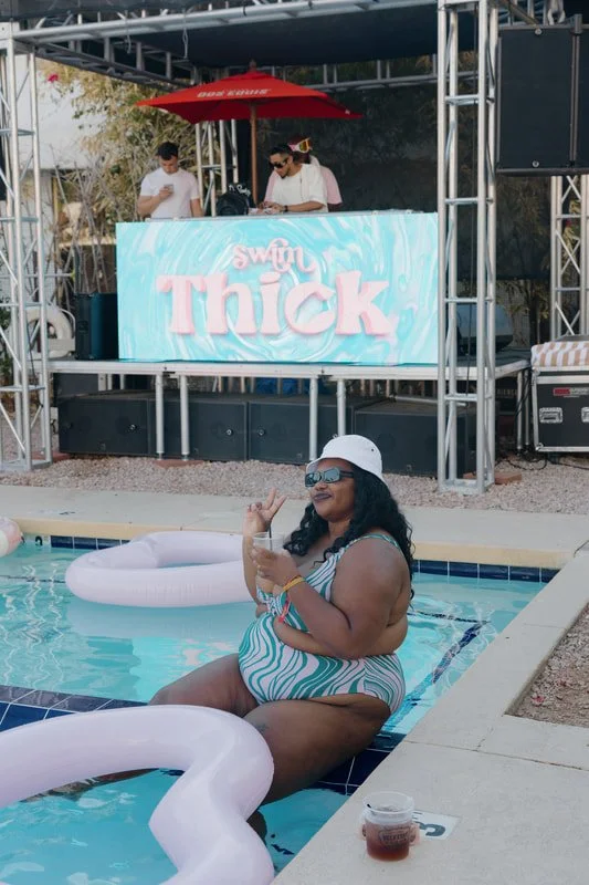 A woman in a striped swimsuit, sunglasses, and a white hat relaxing on the edge of a swimming pool with inflatable pool floats, with a DJ booth and people in the background at a poolside event.