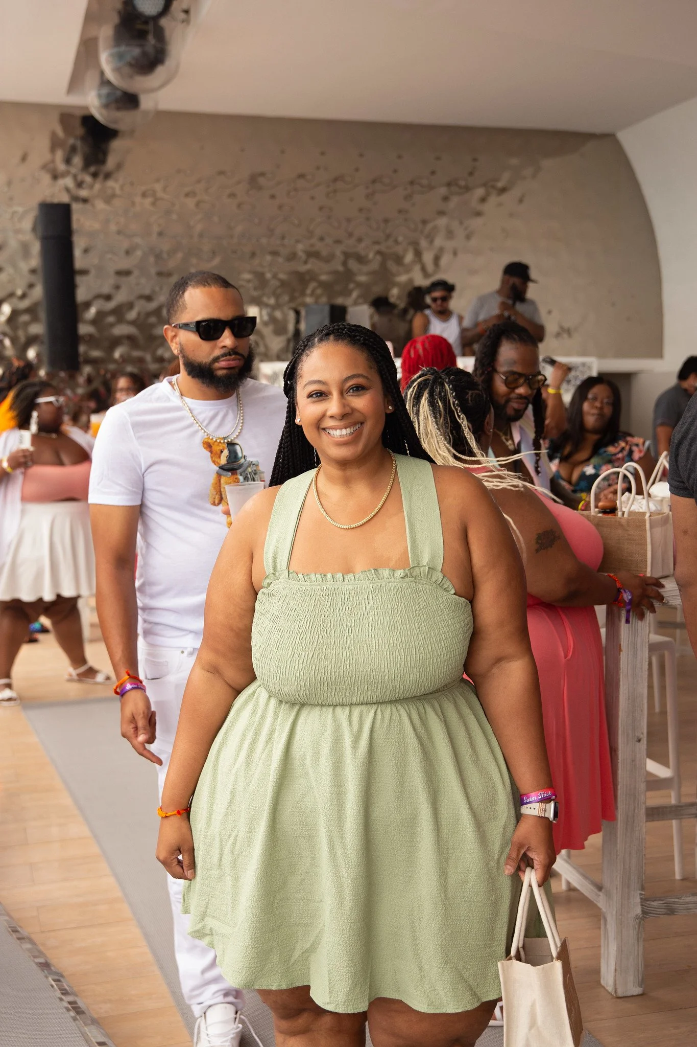 A group of people at a social event indoors, with a woman smiling in the foreground wearing a green dress, and others behind her. The background features a mixed crowd, some wearing sunglasses and casual summer clothing.