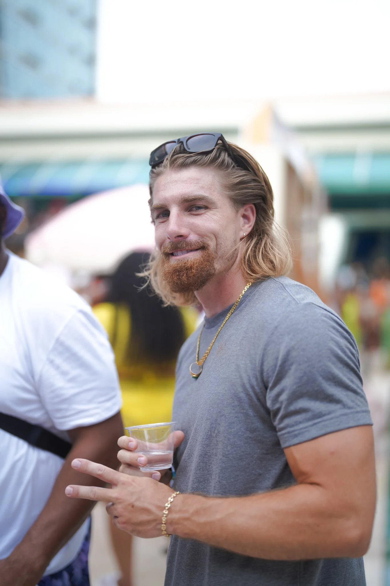 A smiling man with sunglasses on his head, wearing a grey t-shirt and gold jewelry, holding a small glass of clear liquid, standing at an outdoor event with a crowd and colorful tents in the background.