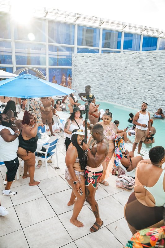 Crowd of people at a pool party with an indoor pool, some people swimming, woman in large hat and bikini, man shirtless holding woman, sunny weather, modern building with glass windows in the background.