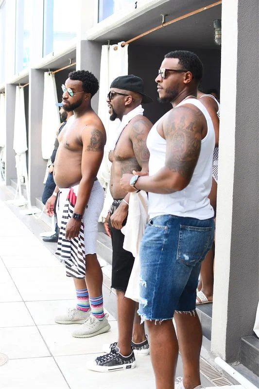 Four men standing in a row outdoors, shirtless or in sleeveless shirts, with sunglasses, next to a poolside cabanas area.