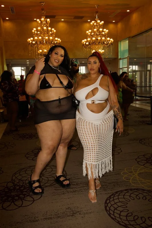 Two women posing together in a hotel lobby with ornate chandeliers overhead. One woman is wearing a black swimsuit with a sheer cover-up, and the other is dressed in a white crochet skirt and cut-out top. There are several people in the background, a