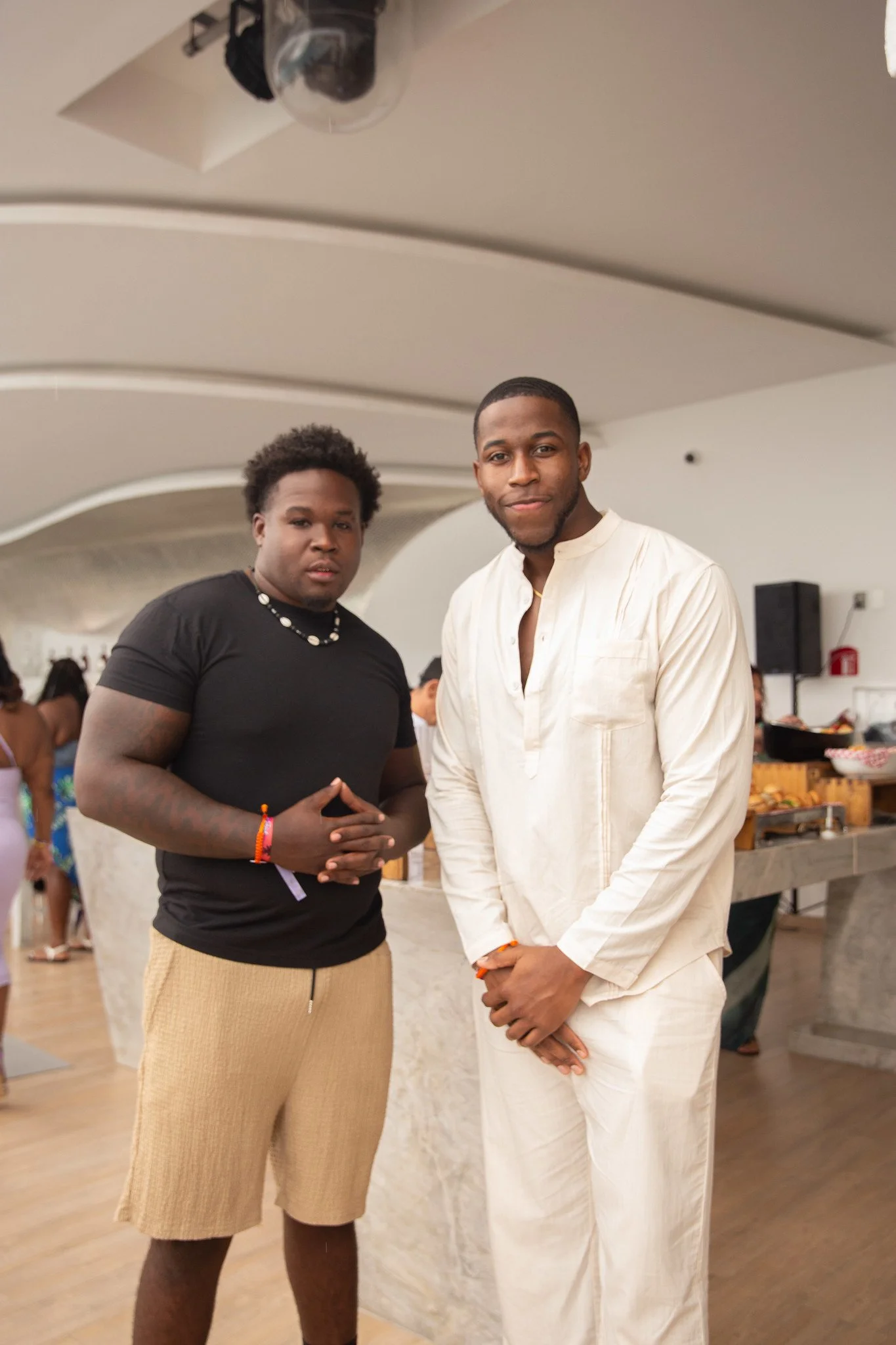 Two young men standing indoors at a social event. One is wearing a black t-shirt and beige shorts, with accessories including bracelets and a necklace. The other is dressed in a cream-colored traditional outfit. They are posing with relaxed expressio