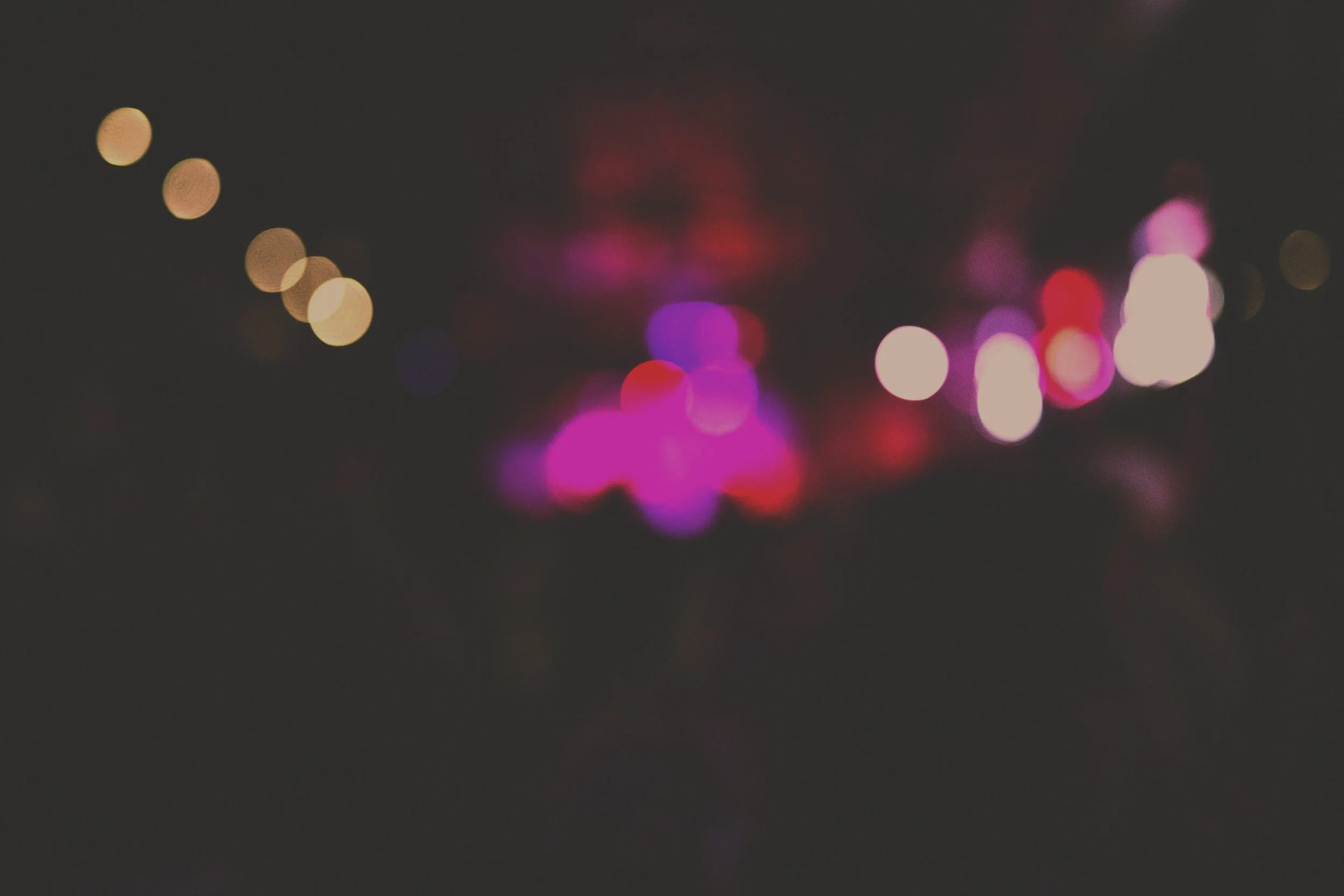 Blurred image of colorful city lights at night, with pink, purple, white, and red lights creating a bokeh effect.