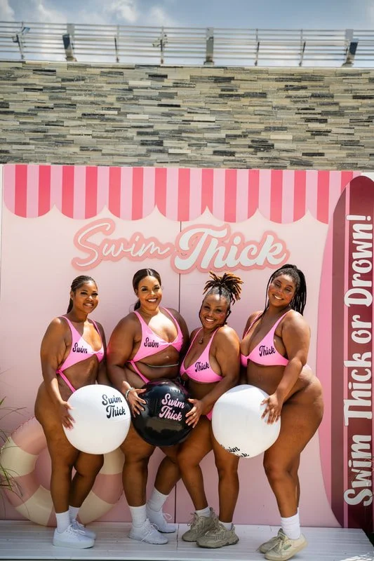 Four women in pink swimsuits with 'Swim Thick' written on them, holding large white and black balls with 'Swim Thick' written on a pink backdrop with a pink striped awning design.