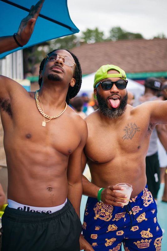 Two shirtless men at an outdoor party, wearing sunglasses and smiling. One has dreadlocks and a gold chain, the other has a beard, a bright yellow cap, and is holding a drink.