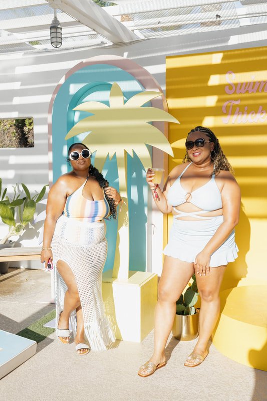 Two women in swimwear standing near a decorative palm tree at a poolside, smiling and holding drinks. The background features a yellow and white striped wall with the words "Summer Trick" partially visible.