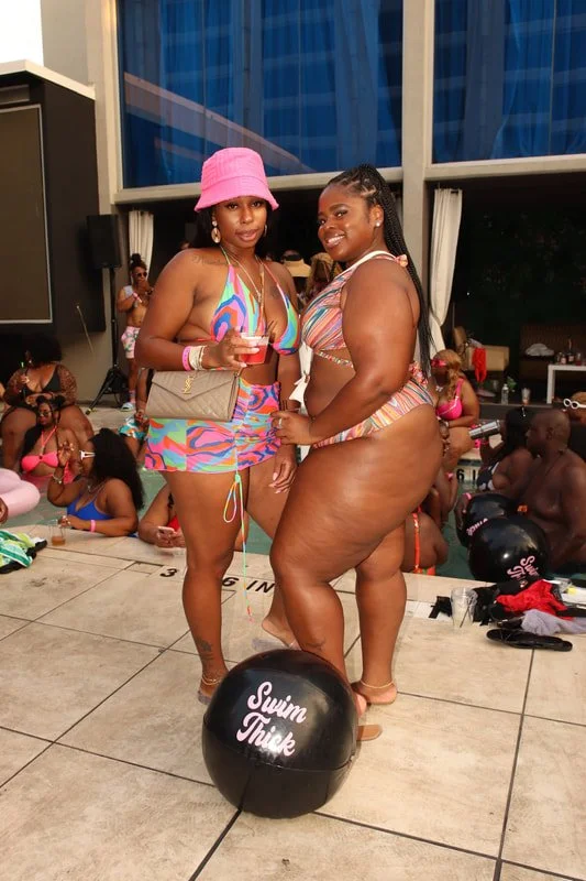 Two women in colorful swimsuits standing by a poolside, one wearing a pink hat and holding a drink, with people in swimsuits and pool accessories in the background.