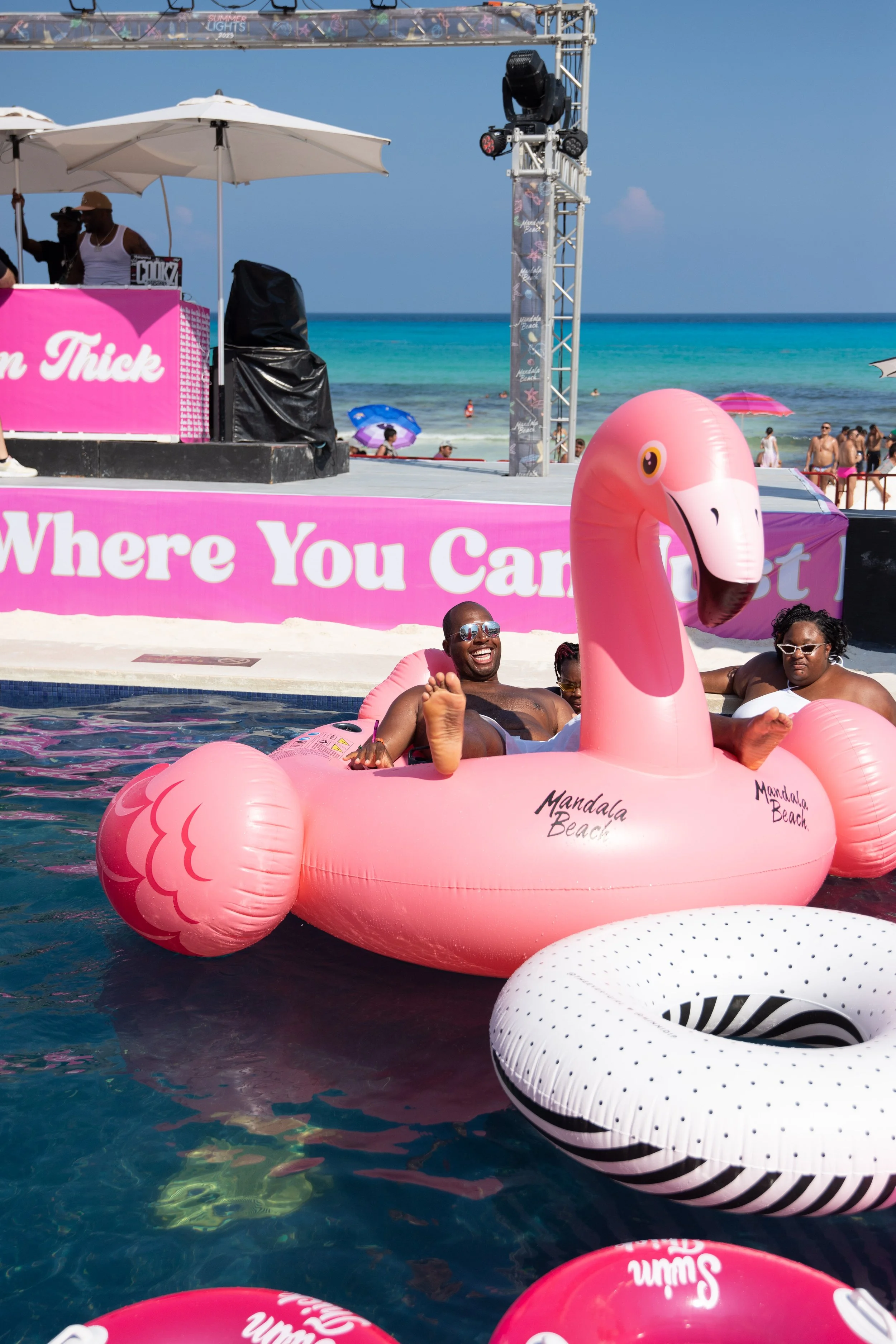 People enjoying a day at the beach on floaties, with a man smiling on a pink flamingo-shaped float and others nearby, at Mandala Beach, with a stage and beachgoers in the background.