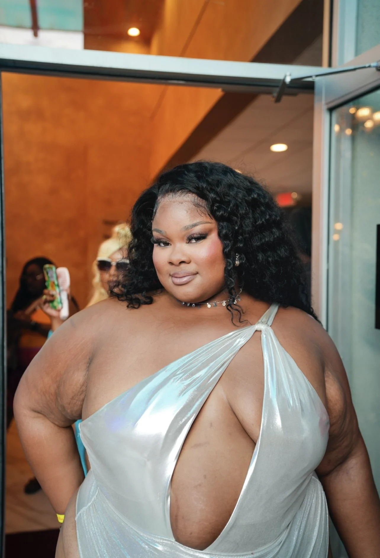 A woman with dark curly hair, wearing a shiny silver dress with a cutout, standing in a doorway.