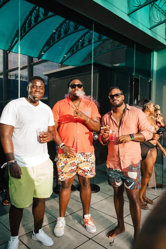 Three men standing outside, dressed casually in summer clothing, enjoying drinks at a social event. Two women are in the background, also at the gathering.