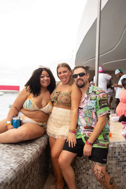 Three friends smiling and posing together outdoors at a poolside party, some wearing swimwear and casual clothes, with a bar and other people in the background.