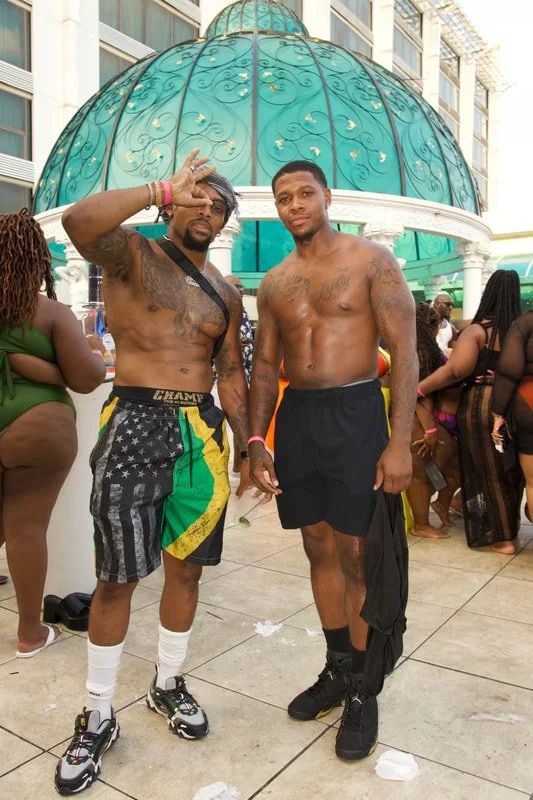 Two shirtless young men standing outdoors, one with gray hair and glasses making a peace sign, the other with short black hair, in front of a domed gazebo at a lively event.