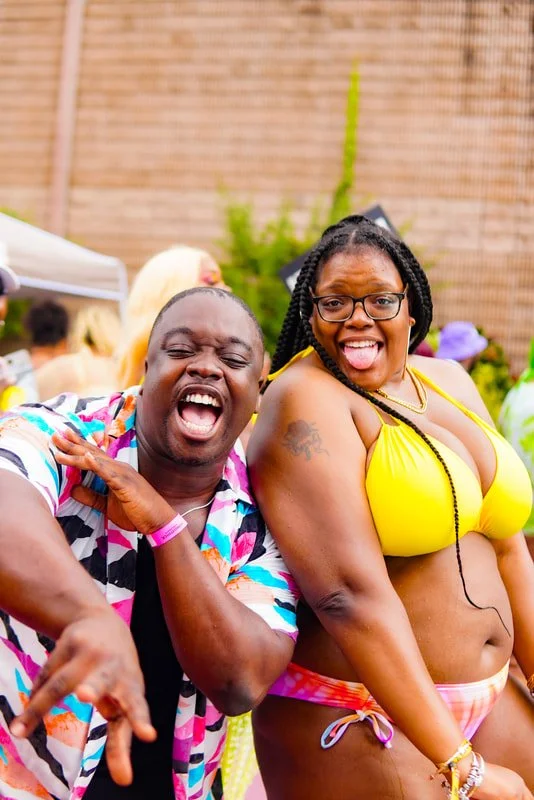 Two people enjoying a lively outdoor party, smiling and making playful gestures. The person on the left is wearing a colorful, striped shirt, and the person on the right is in a yellow bikini top and pink-and-white swim bottoms, with glasses and brai