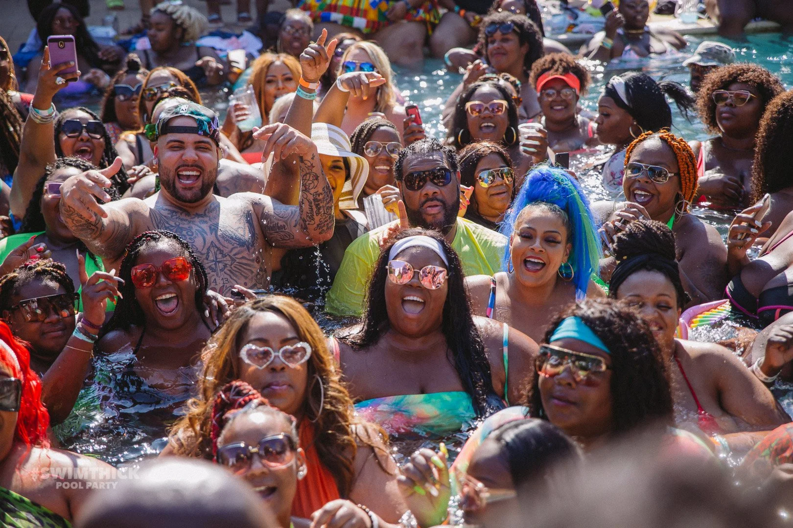 A large group of people in a swimming pool enjoying a fun pool party. They are smiling, laughing, and some are taking photos. The crowd includes diverse individuals wearing colorful swimwear and sunglasses.